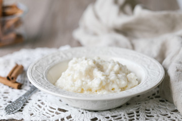 Rice pudding or in a white bowl, on a white lace table cloth. Cinnamons sticks on the table, next to the rice pudding.