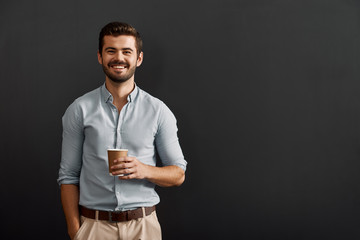 Drinking cappuccino. Cheerful young bearded man holding a cup of hot coffee and looking at camera with smile while standing against dark background