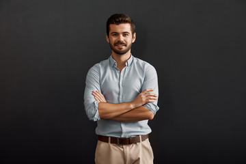 Successful project manager. Cheerful and young bearded man looking at camera with smile and keeping arms crossed while standing against dark background - Powered by Adobe
