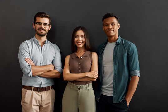 We are great team. Group of three multicultural cheerful young people in casual wear looking at camera with smile while standing against dark background