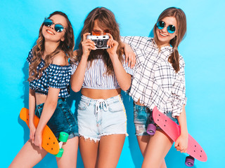 Three sexy beautiful stylish smiling girls with colorful penny skateboards.Women in summer hipster checkered shirt clothes posing near blue wall. Models taking pictures on retro camera