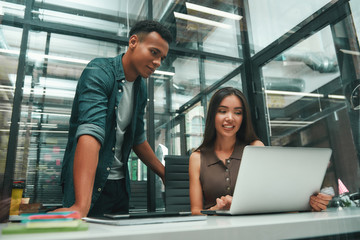 Fototapeta na wymiar Multiracial team. Young asian woman and afro american man in casual wear looking at screen of laptop and discussing new project while working in modern office