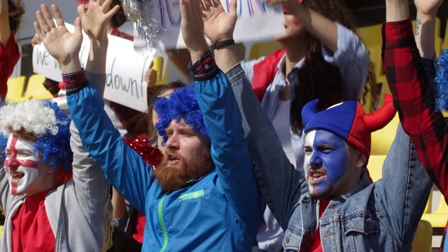 Medium panning shot of loyal male football club fans standing on stadium tribune, clapping hands raised above head, and chanting slogans in unison, and girlfriends with pompoms and banners behind