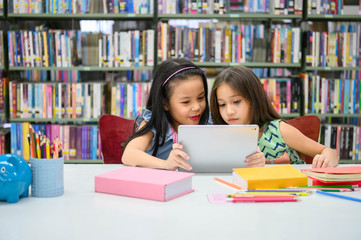 Two little happy cute girls playing on a tablet PC computing device in library at school. Education and self learning wireless technology concept. People lifestyles and friendship. Preschool children