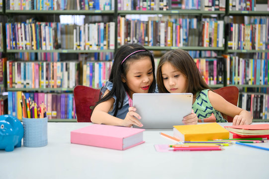Two little happy cute girls playing on a tablet PC computing device in library at school. Education and self learning wireless technology concept. People lifestyles and friendship. Preschool children - Powered by Adobe