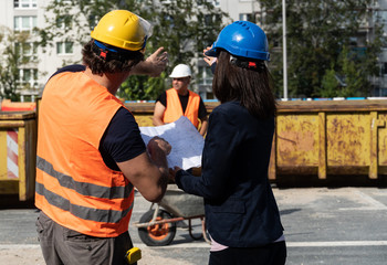Back turned woman civil engineer discussing about plans, blueprints and paperworks with the foreman on construction site. In the background, another building contractor. Selective focus