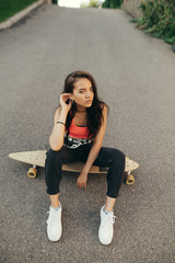 Beautiful girl sits on a skateboard and looks into the camera, wears stylish casual clothes and sneakers, poses on the background of asphalt, top view, street cocnept.