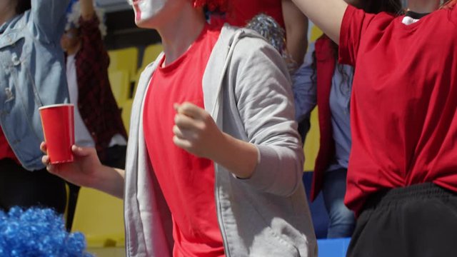 Medium tilting shot of agitated young male fan with painted face, in bright wig, wildly cheering at his team, among fellow supporters with banners, then hurling soda cup and grabbing head in despair