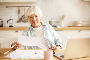 Elderly housewife with gray hair sitting in kitchen with open laptop and papers on table, having...