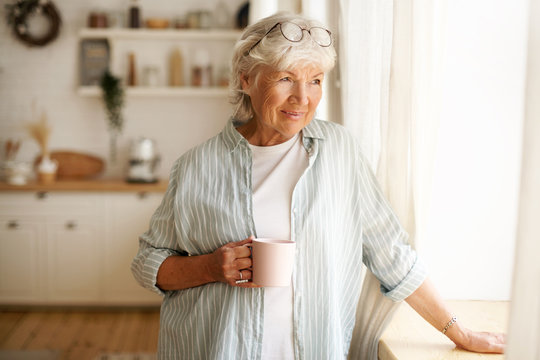 Coziness, domesticity and leisure concept. Portrait of stylish gray haired woman with round spectacles on her head enjoying morning coffee, holding mug, looking outside through window glass
