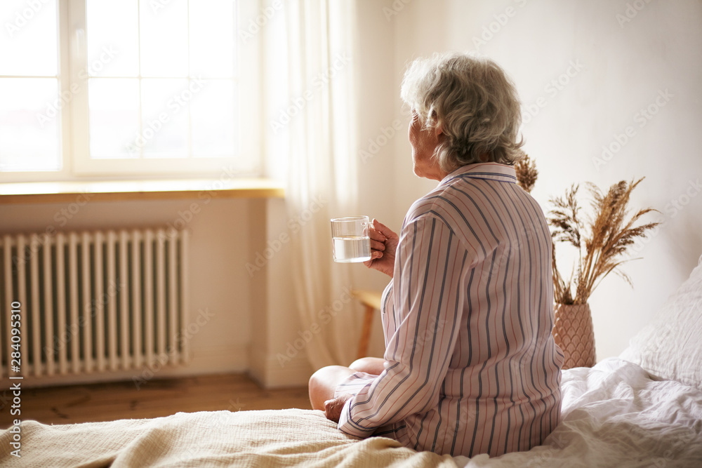 Wall mural rear view of senior sixty year old woman with gray hair holding mug washing down sleeping pill, suff