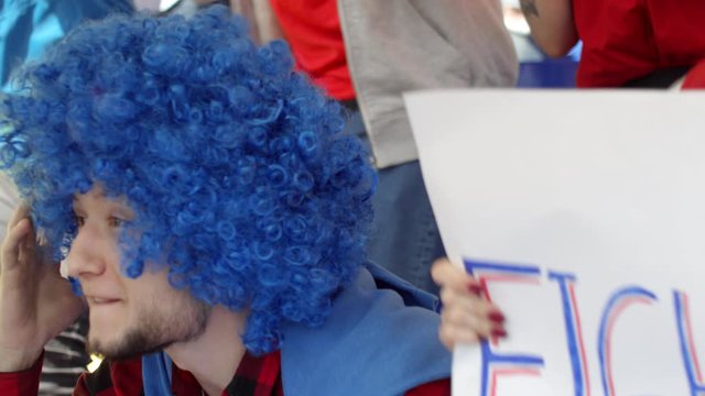 Chest-up shot of 20-something Caucasian male supporter in bright blue fan wig sitting in crowd at stadium, passionately cheering on his team, then expressing dismay and despair at missed goal