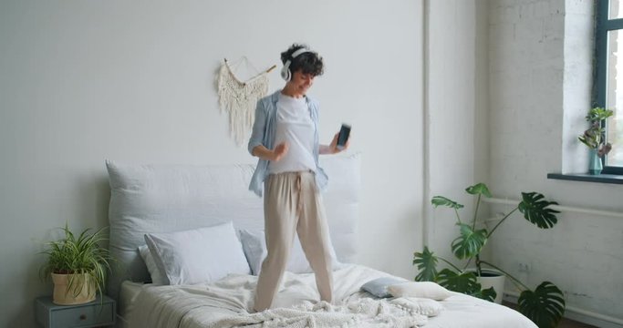 Cute active girl is jumping dancing on bed wearing headphones holding smartphone in hand enjoying leisure time alone in house. Lifestyle and millennials concept.