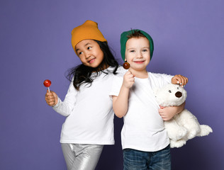 Kids in colorful hats asian girl and caucasian boy with his polar bear toy posing eating lollipop candy on purple