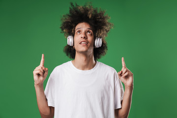 Young curly guy posing isolated over green wall background listening music with headphones pointing to copyspace.