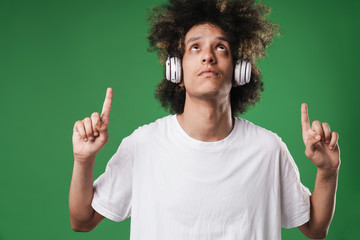 Young curly guy posing isolated over green wall background listening music with headphones pointing...
