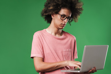 Young serious concentrated curly guy posing isolated over green wall background using laptop...