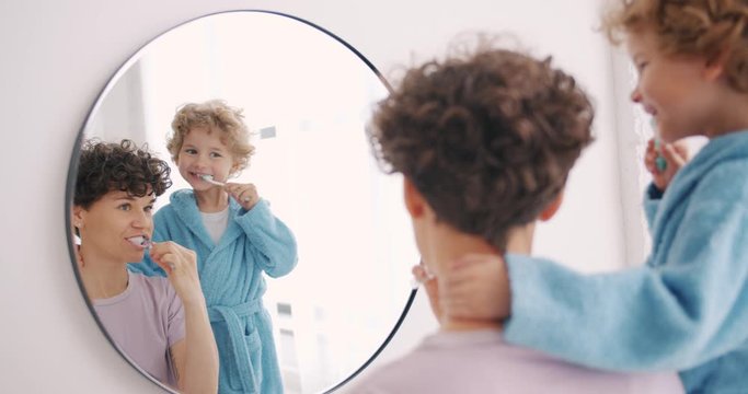 Young woman and small boy are brushing teeth in bathroom looking at mirror smiling using toothbrush and toothpaste. Health care, dental hygiene and people concept.