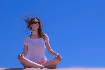 Woman in sunglasses is meditating on vacation. Travel tourist sitting at seashore. Girl meditate looks around sandy beach on blue sky background yoga lotus pose. Traveller tourism in summer wind blow.