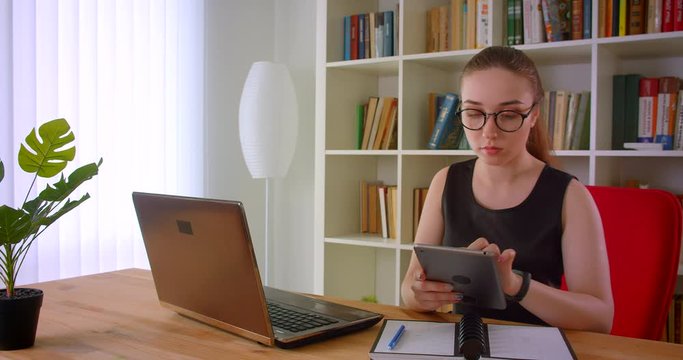 Closeup portrait of young pretty redhead businesswoman in glasses using tablet sitting in front of laptop in office indoors
