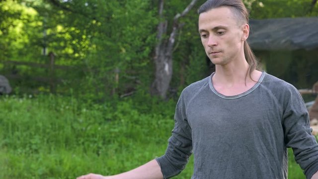 An average plan a young man in Indian clothes and pants Aladdin practices qigong while doing exercises while working with qi energy outdoors in summer next to an old hut