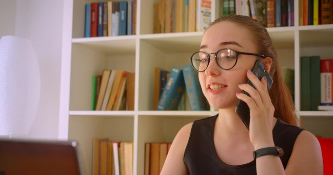 Closeup portrait of young successful redhead businesswoman in glasses using laptop having a phone call talking cheerfully in office indoors