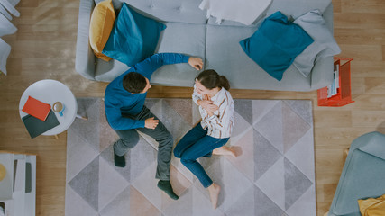 Young Couple is Sitting on a Floor and Talking. They Have an Emotional Dialogue. Cozy Living Room with Modern Interior with Carpet, Sofa, Chair, Table, Book Shelf, Plant and Wooden Floor. Top Down.
