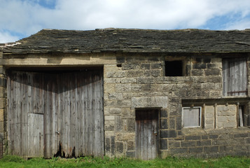 Fototapeta premium ancient abandoned stone barn in a row of rural buildings with empty windows and wooden doors with the pavement overgrown with grass