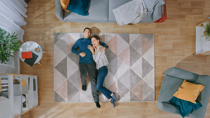 Young Happy Couple is Lying Down on the Floor and Laughing. Man Hugs the Girl. Cozy Living Room...