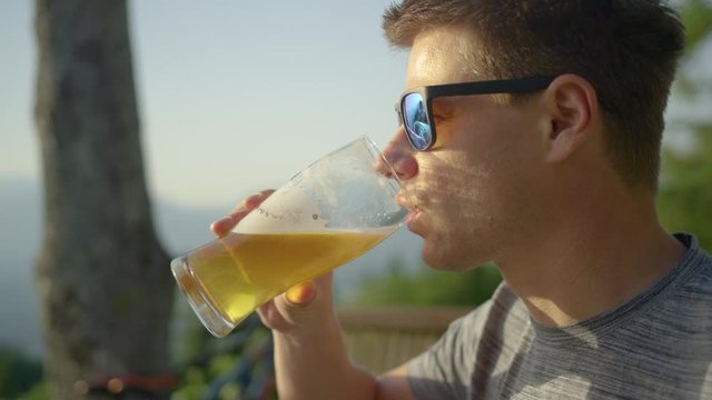 SLOW MOTION, CLOSE UP: Young man wearing sunglasses takes a sip of cold beer on a hot and sunny summer day. Happy Caucasian guy drinks a pint of refreshing lager beer on a beautiful summer afternoon.