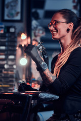 Young attractive tattoo artist in black gloves is tuning her tattoo machine before tattoo session.