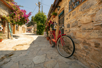 Stone walls of Old Datca and a red bike stands in front of a gift shop. Bougainvillea Begonia flowers in the background.