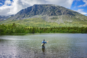 Fototapeta na wymiar Angler beim Fliegenfischen mit Angelroute im Wasser stehend mit Wathose im See vor Bäumen und Berg