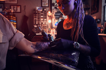 Creative tattoo master with dreadlocks is creating beautiful tattoo on woman's hand at tattoo salon.