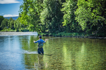 Fototapeta na wymiar Angler beim Fliegenfischen mit Angelroute im Wasser stehend mit Wathose im Fluss vor Bäumen