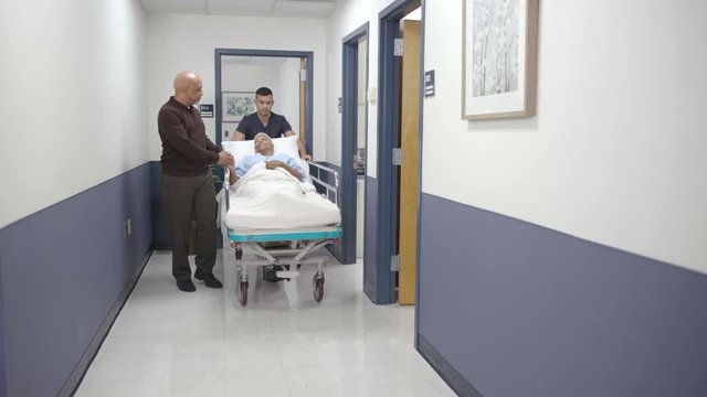 Nurse and husband walk patient down hallway on stretcher camera right left
