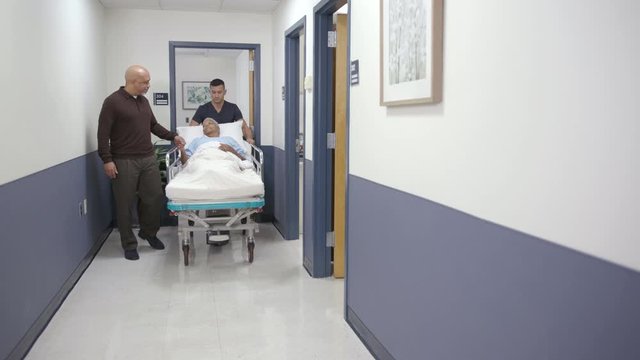 Nurse and husband walk patient down hallway on stretcher slight camera move