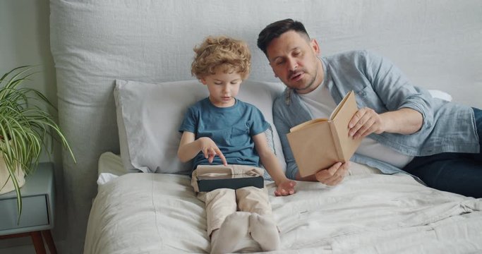 Loving father middle-aged man is reading book to cute child while kid is using tablet touching screen in bed in apartment. Family, literature and gadgets concept.