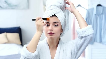 woman in bathrobe applying makeup foundation with cosmetic brush