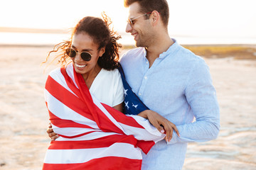 Photo of attractive multiethnic couple wrapped in american flag smiling and hugging together by seaside