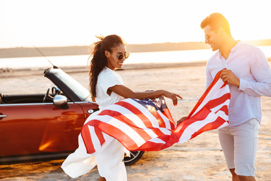 Photo of romantic multiethnic couple smiling and holding american flag while walking by car outdoors - Powered by Adobe