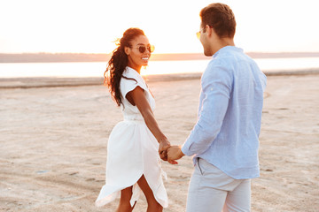 Image of stylish multiethnic couple smiling and holding hands together while walking by seaside at sunset