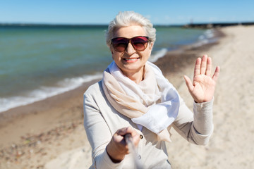 old people and leisure concept - happy smiling senior woman in sunglasses taking picture by selfie stick and waving hand on beach in estonia