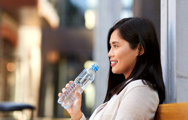 lifestyle and people concept - happy smiling young asian woman drinking water flom plastic bottle sitting on wooden city bench