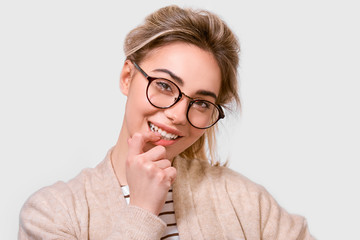 Beautiful positive female dressed in casual outfit, wearing round transparent eyeglasses with pleasant smile, looking to the camera, making flirt gesture and posing over white wall. People emotions