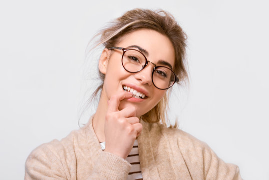 Portrait of beautiful positive female dressed in casual outfit, wearing round transparent eyeglasses with pleasant smile, looking to the camera and posing over white wall. People emotions