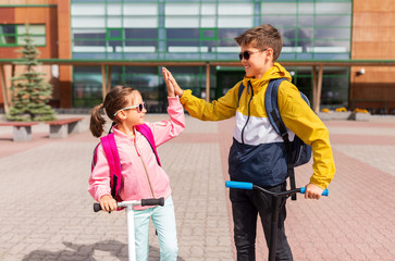 education, childhood and people concept - happy school children with backpacks riding scooters and making high five outdoors
