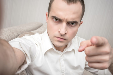 Attractive young man in white shirt makes a video call and points his finger at the camera, sitting...