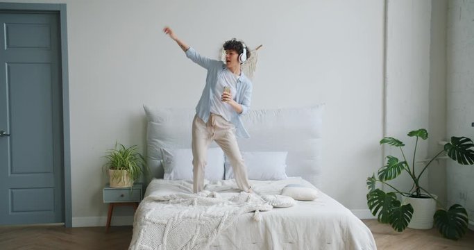 Slow motion of happy young woman dancing on bed wearing headphones having fun listening to music holding smartphone. Youth lifestyle and culture concept.
