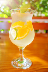 Natural citrus lemonade with slices of orange, lemon and lime in a glass on a wooden table against the background of a gazebo and bushes. Vitamin drinks concept. Bright sunshine.